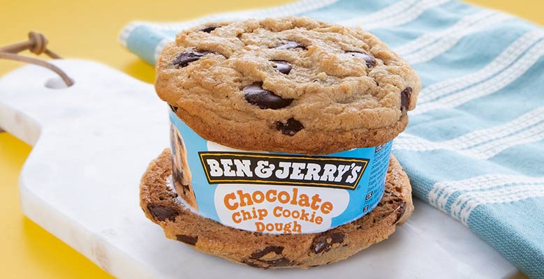 Ice cream sandwich made with two chocolate chip cookies and Ben & Jerry's Chocolate Chip Cookie Dough ice cream sits on a cutting board on a yellow background.
